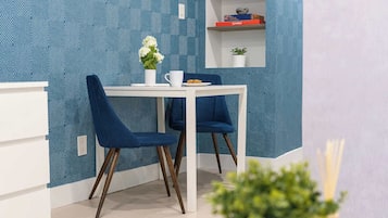 A dining area with a white table and two blue chairs, a vase with flowers, and a shelf with books and a plant.