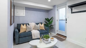 Un salon moderne avec un canapé bleu foncé, des coussins blancs, une table basse ronde ornée de livres et d’une plante, et une grande fenêtre.