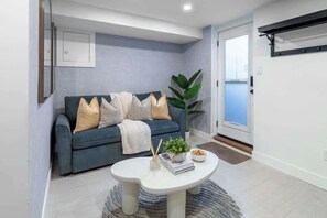 A modern living room with a dark blue sofa, white cushions, a round coffee table with books and a plant, and a large window. - Modern Basement Suite Heart of Ossington (Toronto)