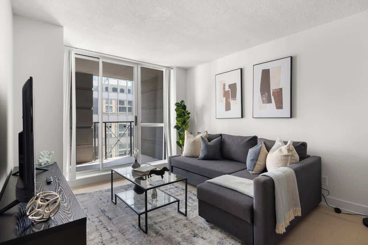 Un salon moderne avec un canapé gris, une table basse en verre et un téléviseur à écran plat. Une grande fenêtre offre une vue sur les bâtiments environnants.