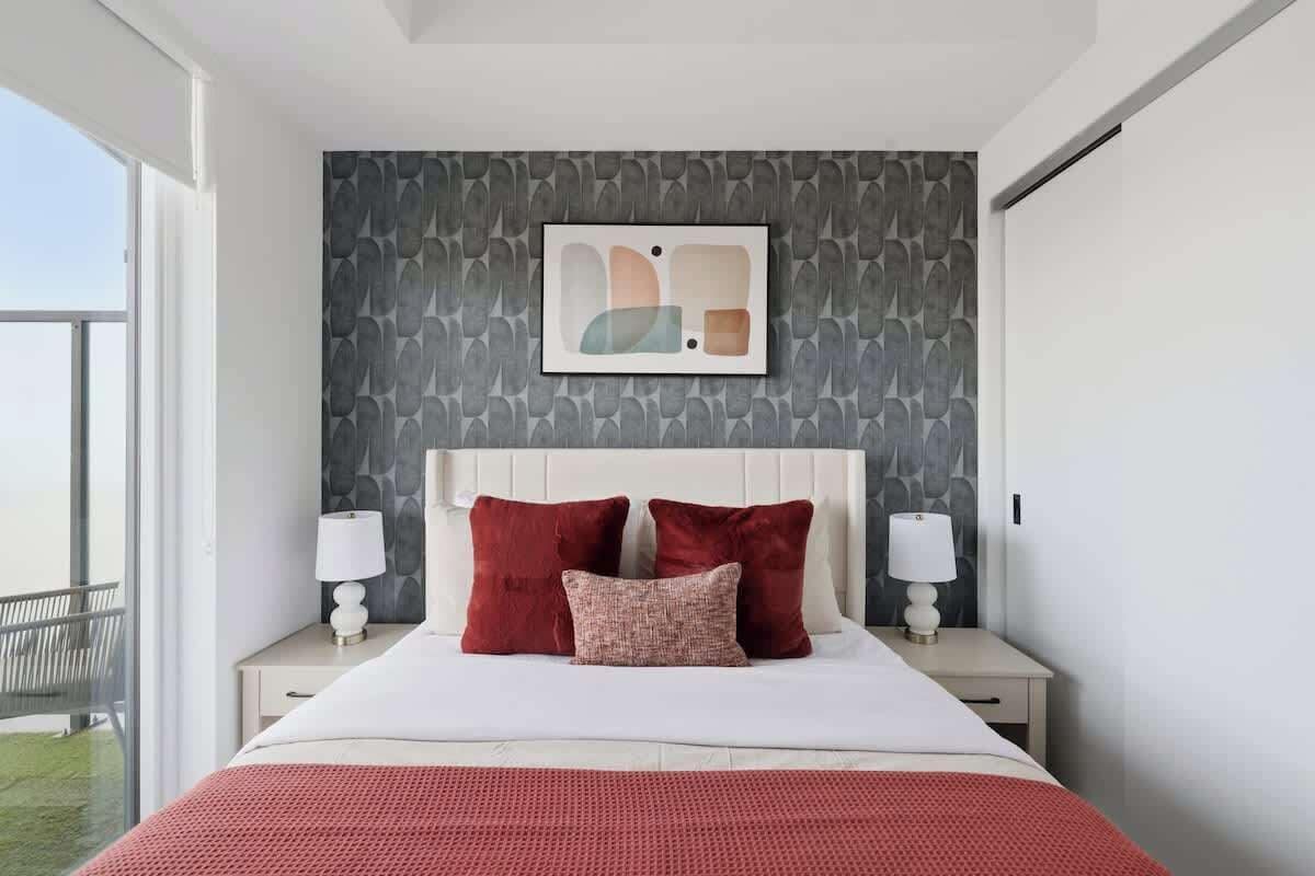 A modern hotel room with a large bed, two nightstands, a wall-mounted artwork, and a view of the outdoors.