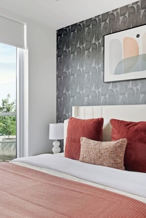 Ein Schlafzimmer mit einem großen Bett, einem weißen Kopfteil und Blick ins Freie durch ein Fenster.