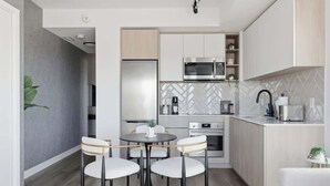 Una cocina moderna con armarios blancos, un refrigerador de acero inoxidable, un microondas y una cafetera. Hay una mesa de comedor con cuatro sillas.