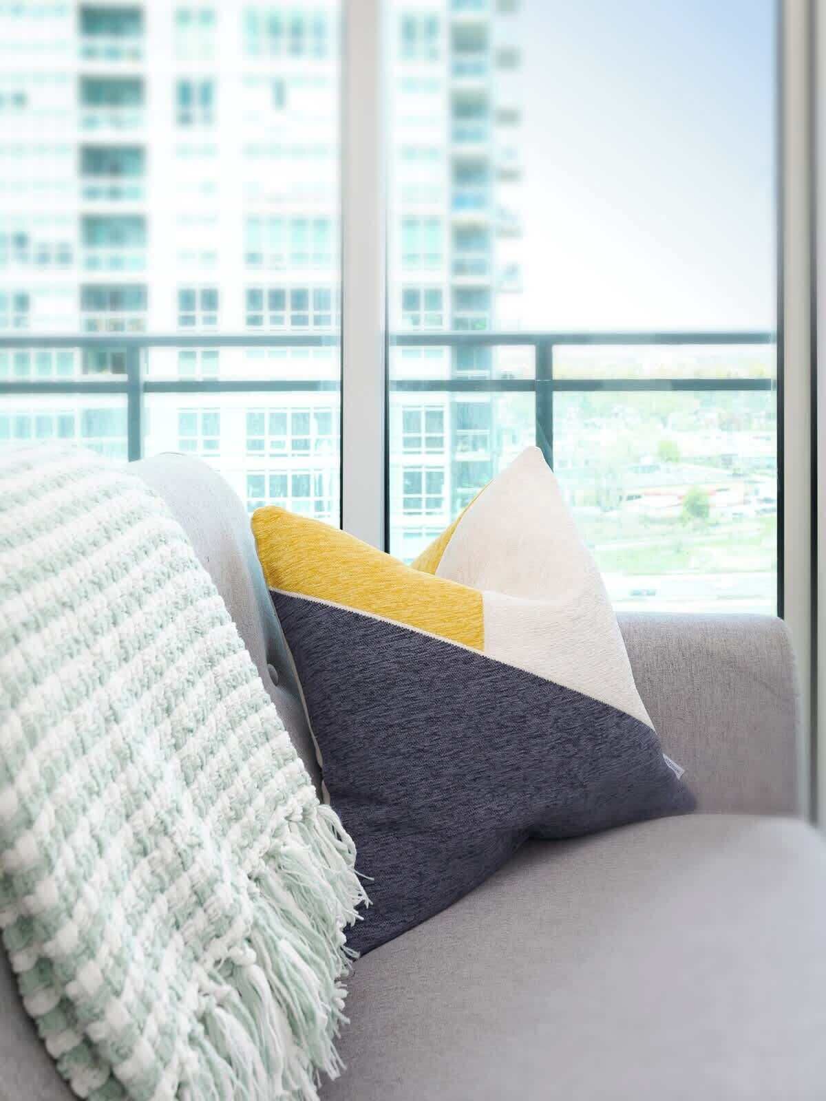 A sofa with a throw blanket and pillows, a window with a view of buildings, and a balcony railing.