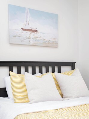 Ein Bett mit gelb-weiß gestreiften Kissen, einer weißen Tagesdecke und einem Bild eines Segelboots an der Wand.