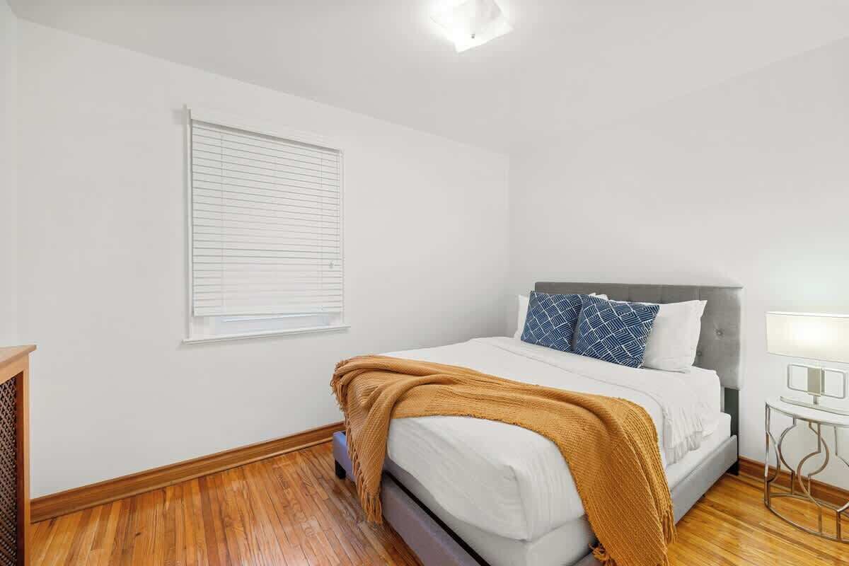 A bedroom with a bed, a bedside table, a lamp, and a window with blinds.