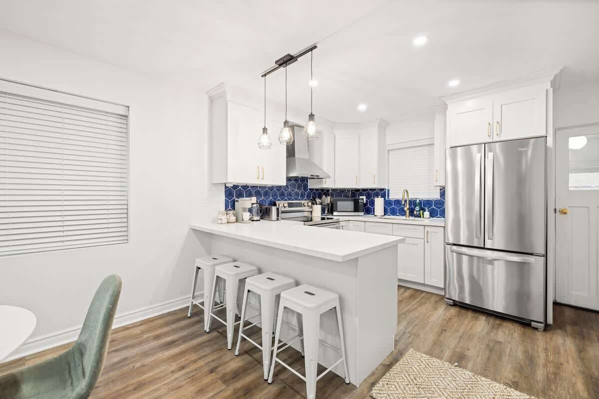 A modern kitchen with a central island, bar stools, stainless steel appliances, and a blue-tiled backsplash.