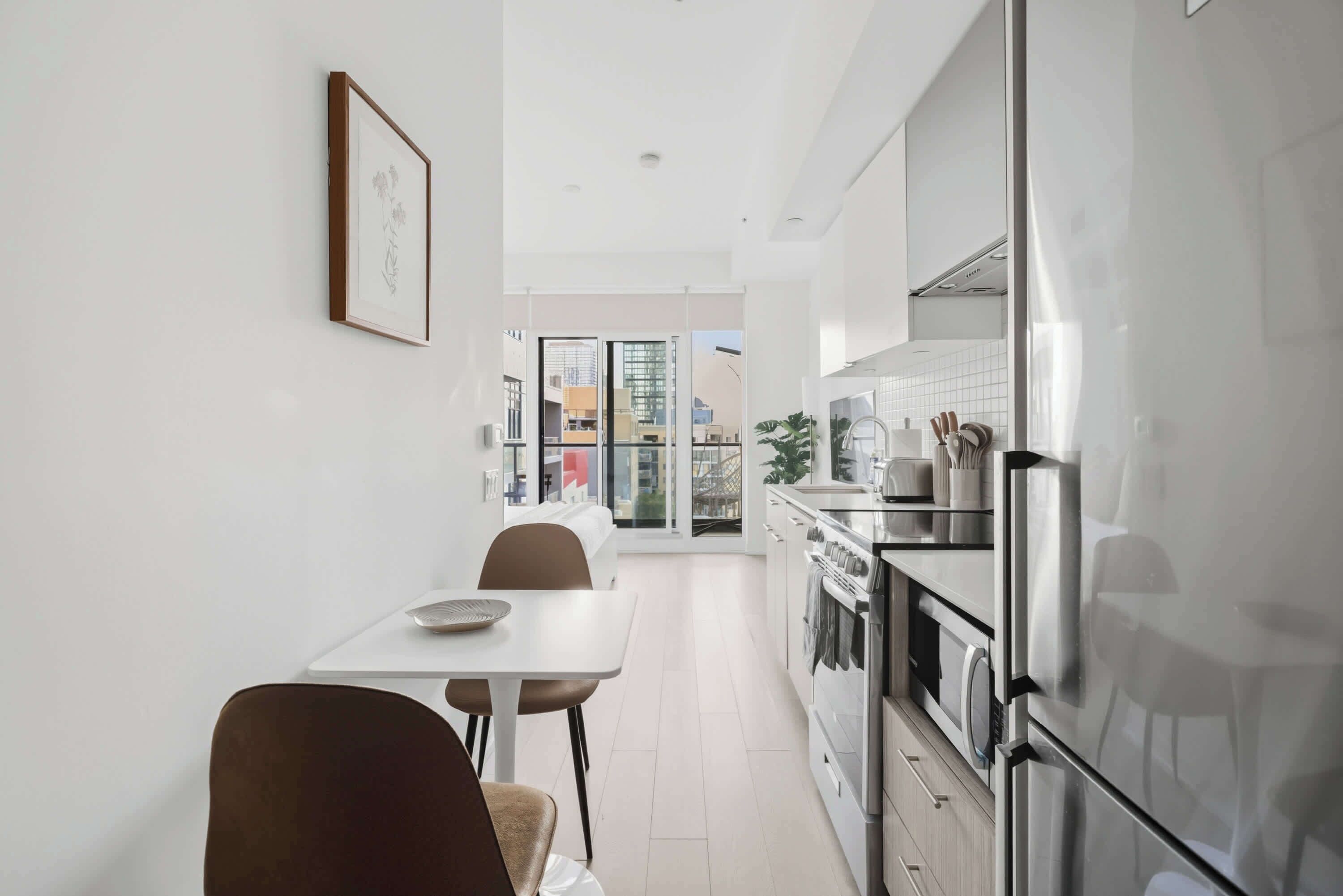 A modern kitchen with stainless steel appliances, a dining area with a white table and chairs, and a view of the city through large windows.