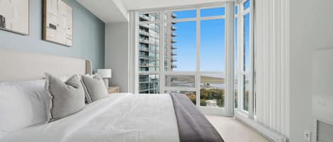 A bedroom with a large bed, a view of a building through a glass door, and a window with a view of the sky.