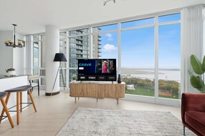 A modern living room with a large TV displaying 'CRAVE' on the screen, a dining area with a white table and chairs, and a view of a cityscape through large windows.