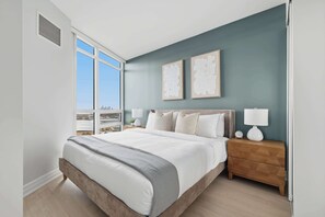 Un dormitorio moderno con una cama grande, una mesita de noche, dos lámparas de pared y vistas a la ciudad a través de un ventanal amplio.