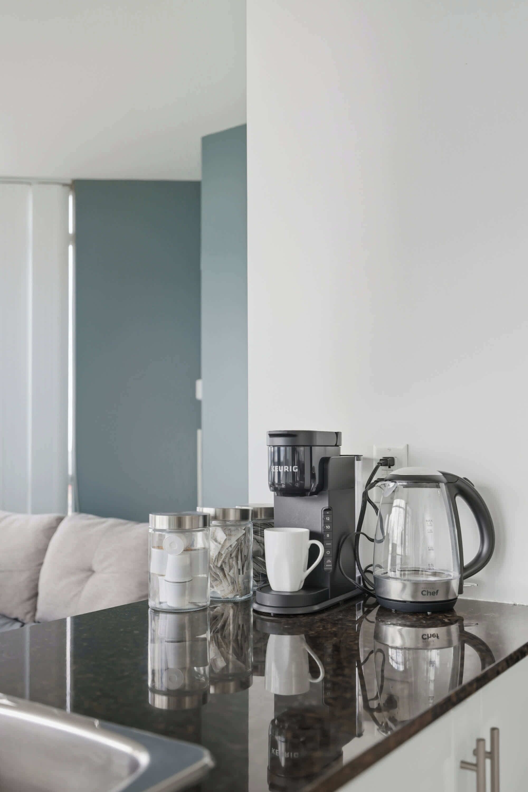 Een moderne hotelkamer met een zwarte koffiezetapparaat, een glazen koffiekan en een witte mok op een reflecterend oppervlak.