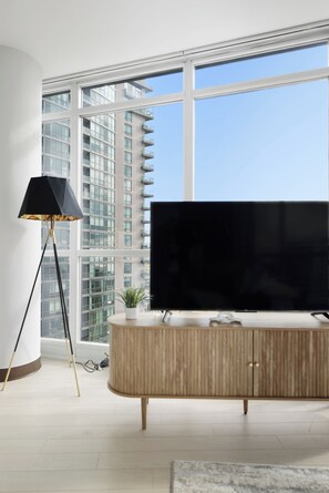 A modern living room with a large flat-screen TV, a floor lamp, and a view of a high-rise building through large windows.