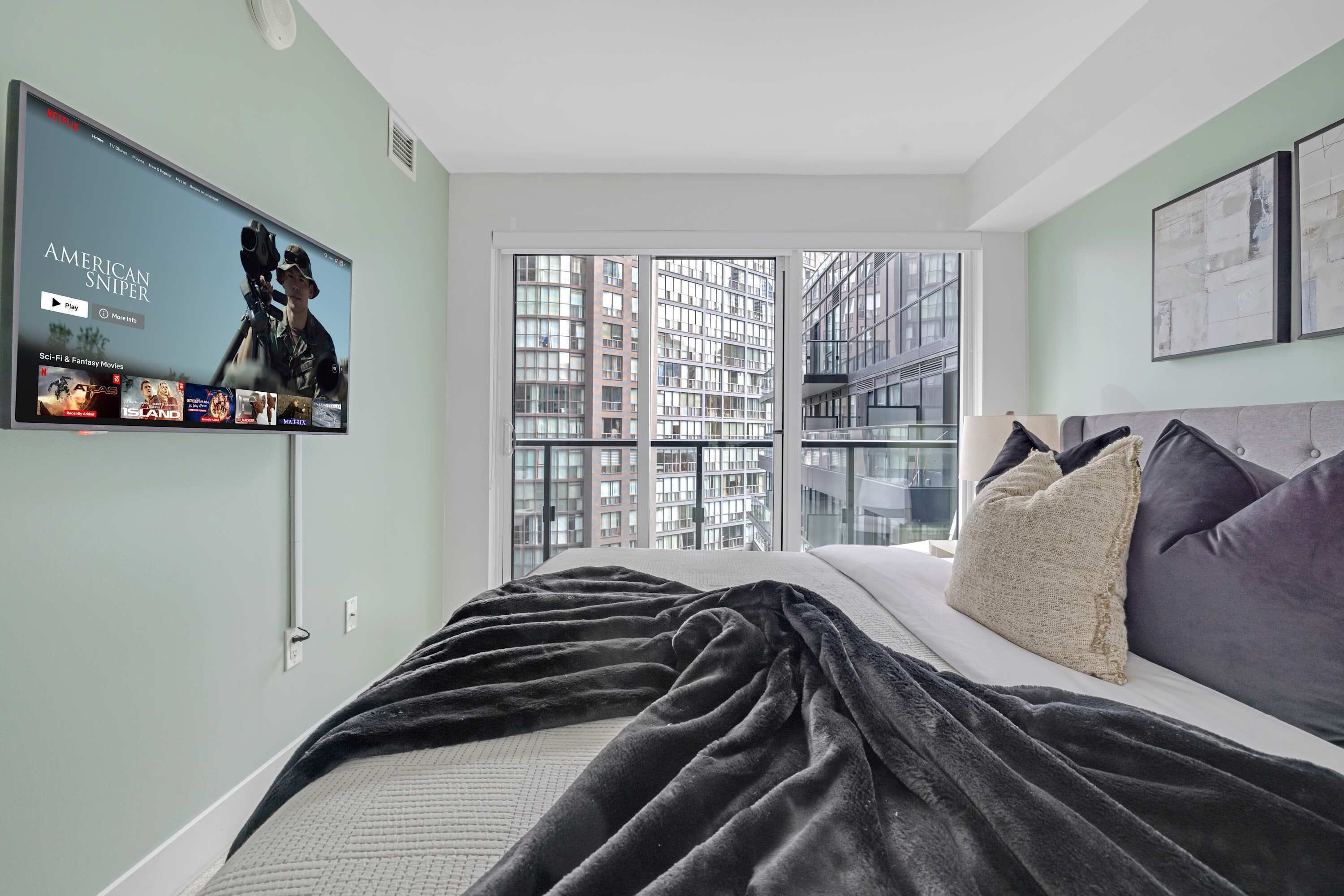 Un dormitorio moderno con una cama grande, un televisor en la pared y vistas de un paisaje urbano a través de la ventana.