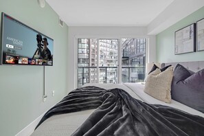 A modern bedroom with a large bed, a television on the wall, and a view of a cityscape through the window. - Financial District 2BR Condo, 55" TV, Free Parking (Toronto)