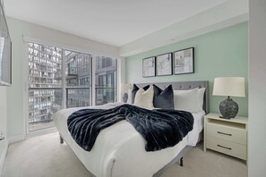 Une chambre moderne avec un grand lit, une table de chevet, une lampe et une vue sur le paysage urbain grâce à de grandes fenêtres du sol au plafond.