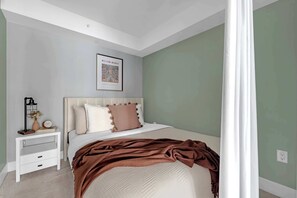 Ein Schlafzimmer mit einem Bett, einem Nachttisch, einem gerahmten Bild und einem weißen Vorhang.