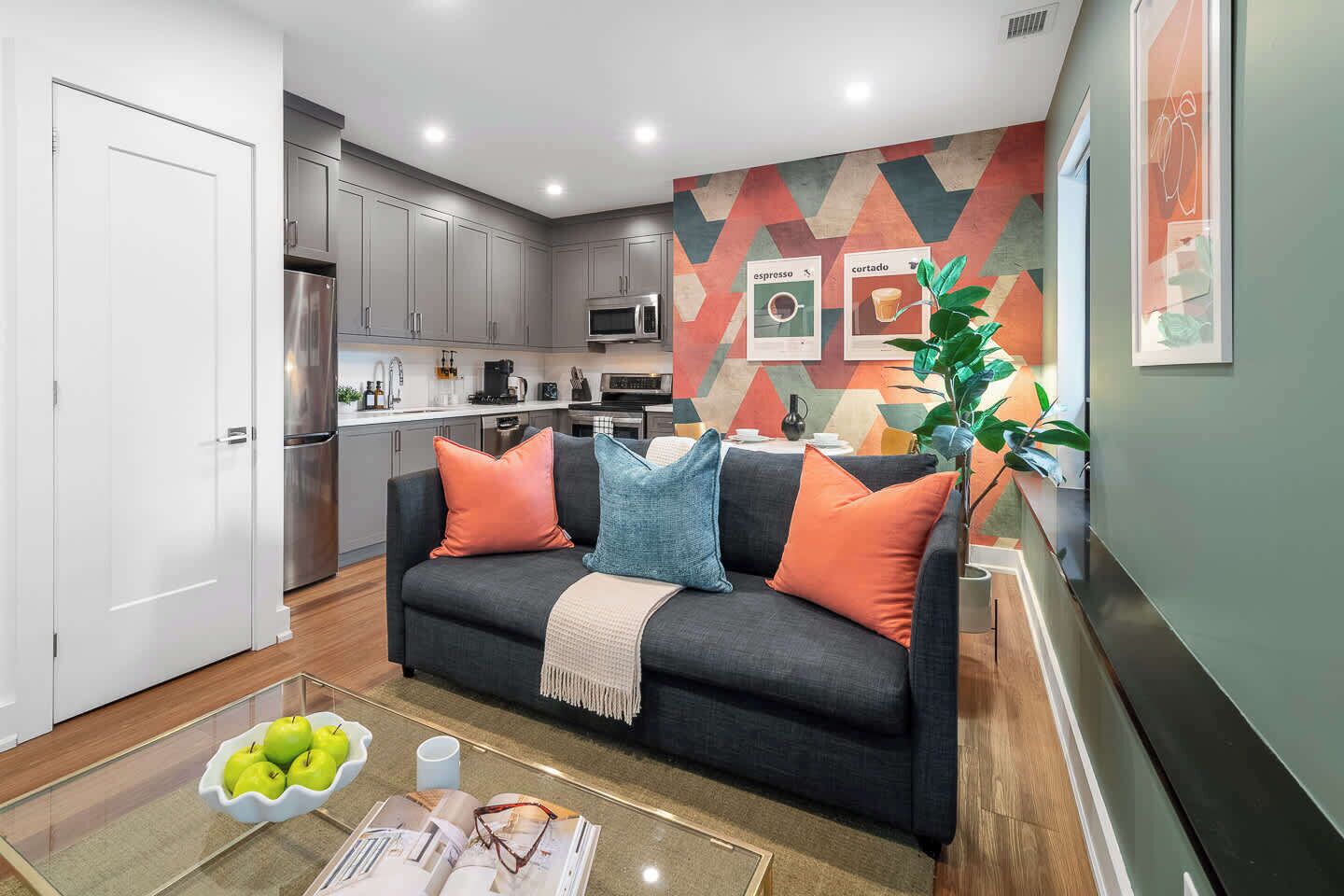 A modern living room with a grey sofa, a coffee table with apples and glasses, and a kitchen area with a microwave and cabinets.