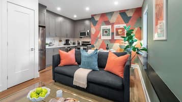 A modern living room with a grey sofa, a coffee table with apples and glasses, and a kitchen area with a microwave and cabinets.