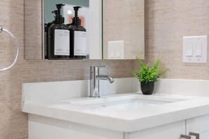 Ein Badezimmer mit einem weißen Waschbecken, einem modernen Wasserhahn, einem Spiegel über dem Waschbecken und einer Topfpflanze auf der Arbeitsfläche.