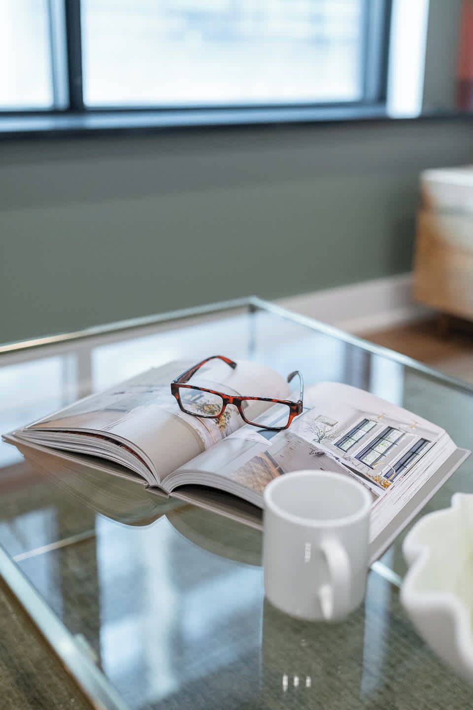 Un magazine, une paire de lunettes posées dessus, une tasse blanche et une table en verre dans une pièce avec une fenêtre.
