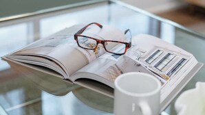 A magazine with glasses on top, a white mug, and a glass table in a room with a window.