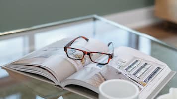 Un magazine, une paire de lunettes posées dessus, une tasse blanche et une table en verre dans une pièce avec une fenêtre.