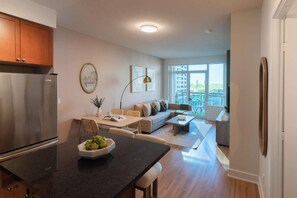 A modern kitchen with a stainless steel refrigerator, a dining area with a table and chairs, a living room with a sofa and a coffee table, and a large window offering a view of the city. - Elegant 2BR Condo in Prime Downtown Location | Parking Available (Toronto)