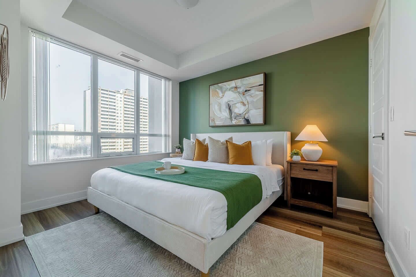 A bedroom with a large bed, a bedside table with a lamp, and a view of the city through large windows.
