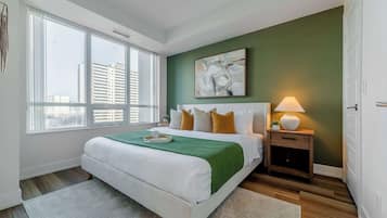 A bedroom with a large bed, a bedside table with a lamp, and a view of the city through large windows.