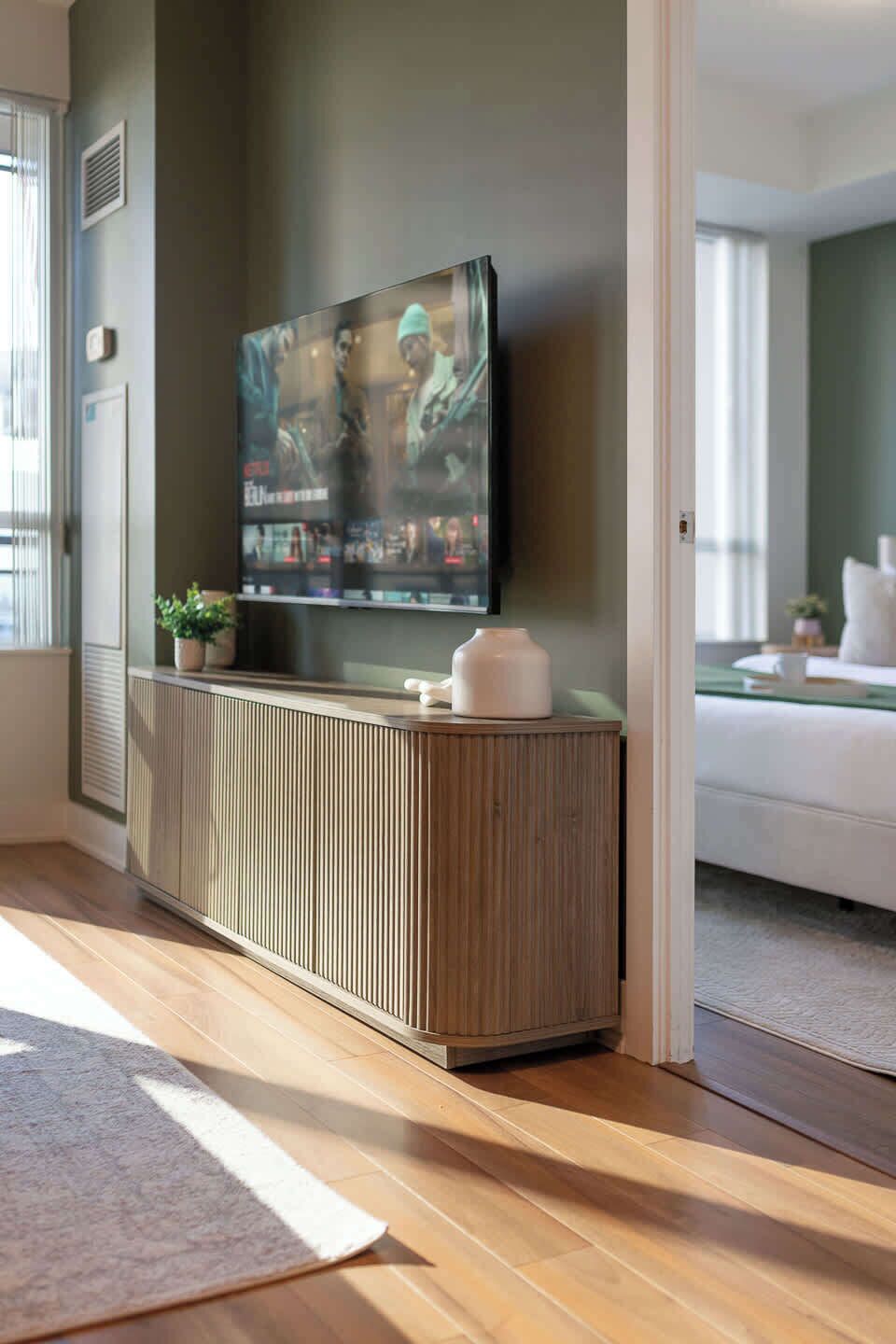 A modern living room with a large flat-screen TV mounted on a wooden cabinet, a white sofa, and a small potted plant.