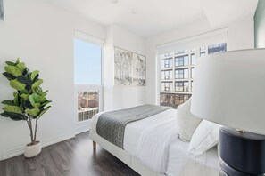 A modern bedroom with a large bed, a potted plant, and a city view. - Downtown Toronto 1BR | Skyline Balcony (Toronto)