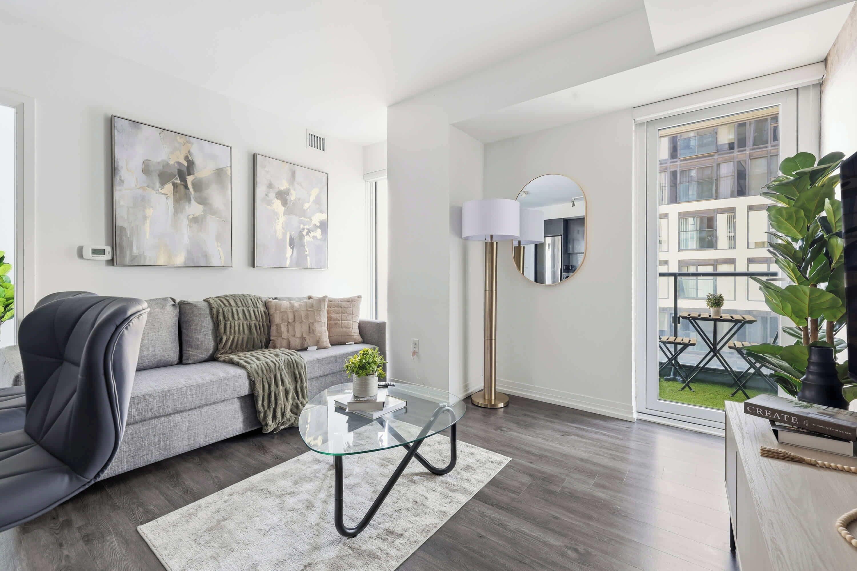 A modern living room with a grey sofa, abstract wall art, a glass coffee table, a floor lamp, and a view of a balcony with plants.