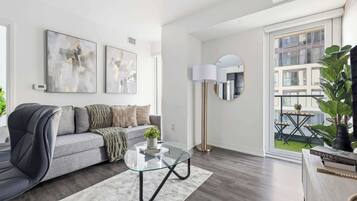A modern living room with a grey sofa, abstract wall art, a glass coffee table, a floor lamp, and a view of a balcony with plants.