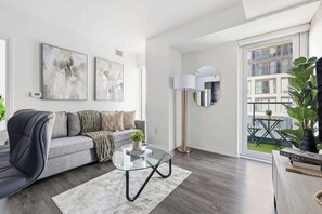 A modern living room with a grey sofa, abstract wall art, a glass coffee table, a floor lamp, and a view of a balcony with plants.