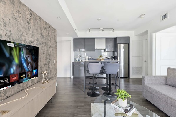 A modern living room with a flat-screen TV, a sofa, a coffee table with books and a remote, and a kitchen in the background.