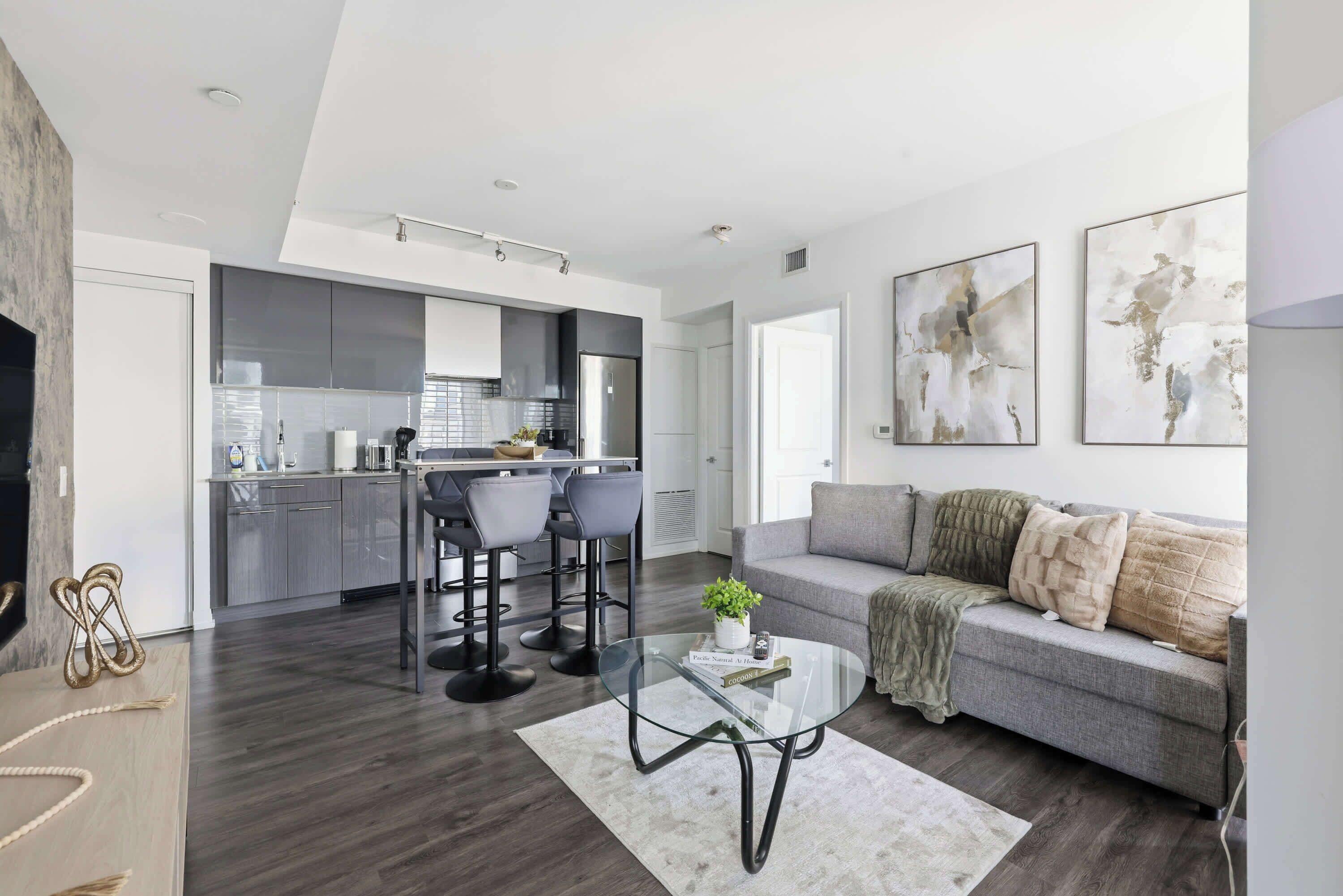 A modern living room with a grey sofa, a glass coffee table, and a kitchen area with grey cabinets and a stainless steel refrigerator.