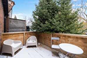 A wooden deck with a snow-covered floor, a large evergreen tree, and two white chairs and a small table. - Parkdale 1BR Hideaway | Patio & Free Parking (Toronto)