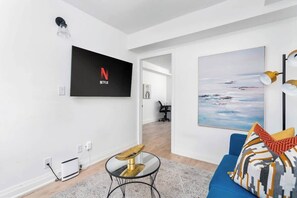A modern living room with a flat-screen TV mounted on the wall, a blue sofa with patterned cushions, a glass coffee table, and a painting of a landscape.