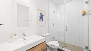 Un baño moderno con lavamanos blanco, tocador de madera, inodoro y cabina de ducha de vidrio.