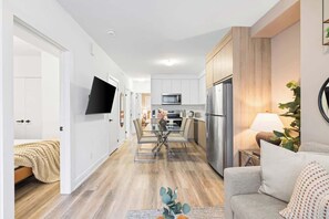 A modern kitchen with stainless steel appliances, a dining area with a glass table, and a living space with a sofa and a plant. - Spacious Bloordale Condo | Quick Transit Access (Toronto)
