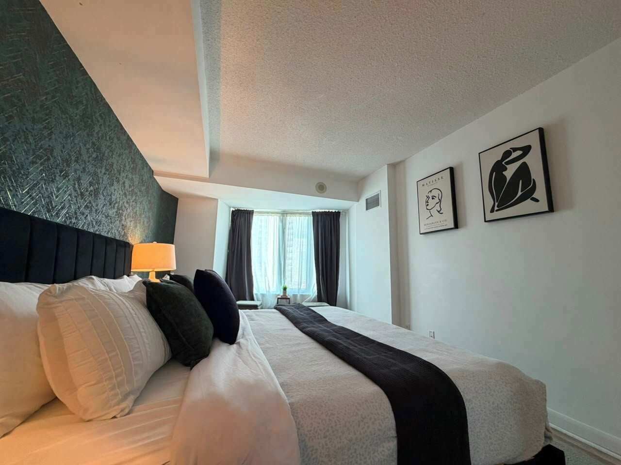 A hotel room with a bed, a nightstand, a lamp, a window with curtains, and two framed pictures on the wall.