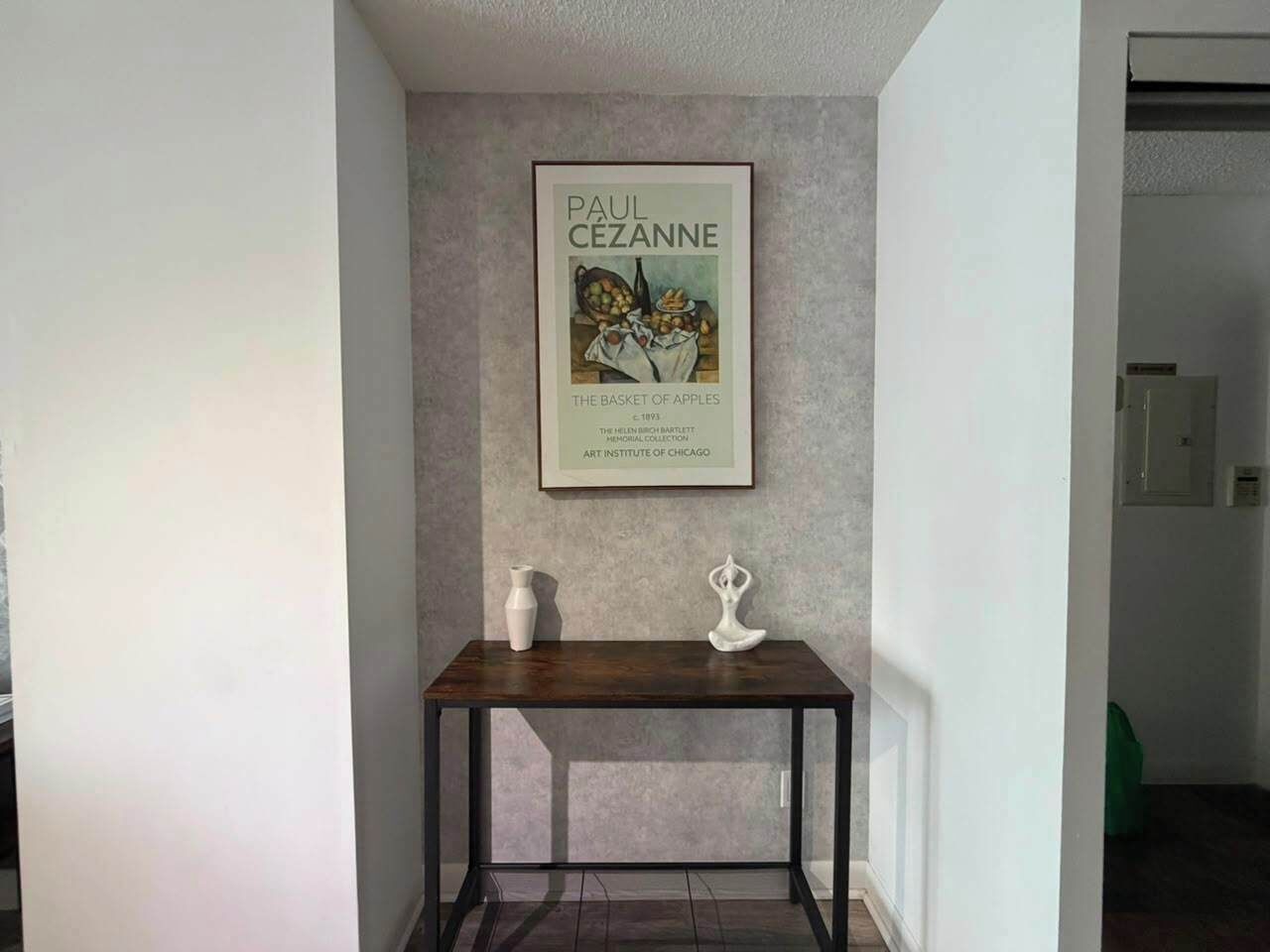 A framed artwork of Paul Cezanne's 'The Basket of Apples' on a wall above a dark wooden table with two white vases.