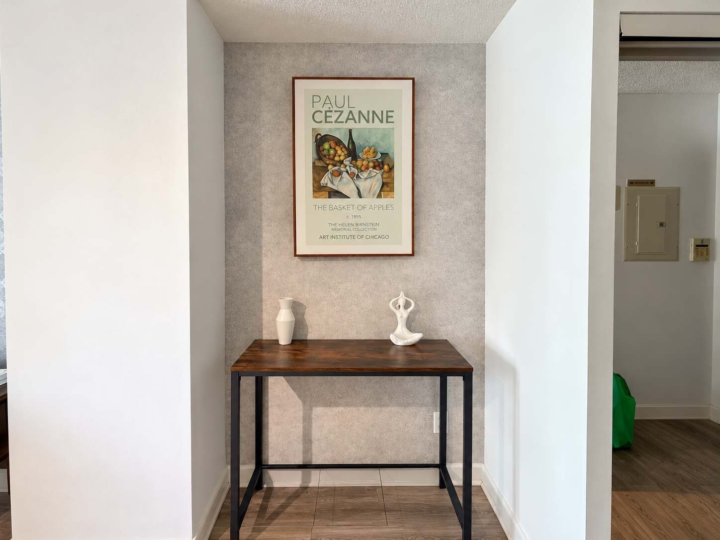 A framed artwork titled 'The Basket of Apples' by Paul Cezanne, displayed on a wall above a small table with a vase and a sculpture.