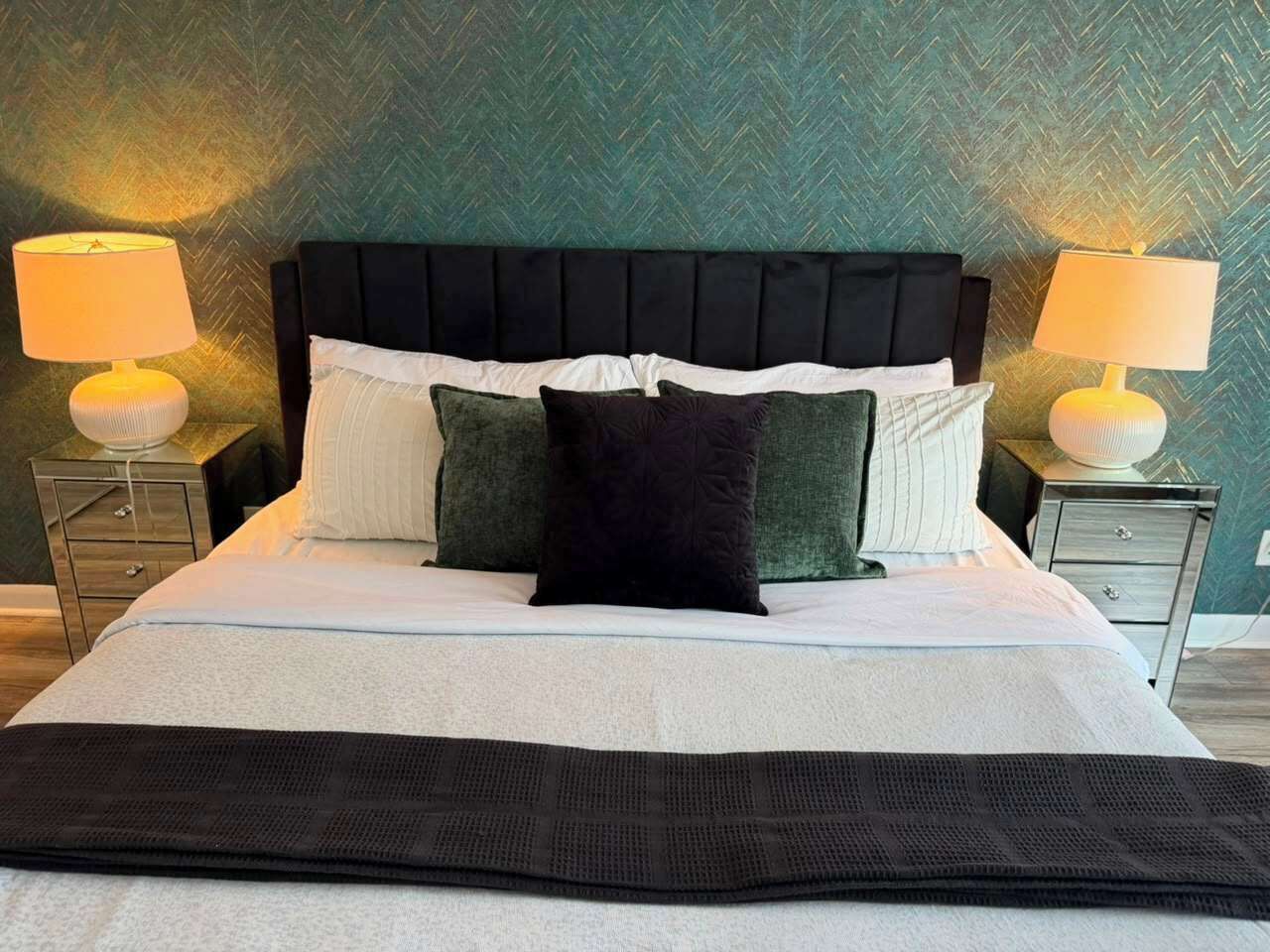 A bed with a black headboard, white and green pillows, and a black throw blanket. There are two bedside tables with lamps.