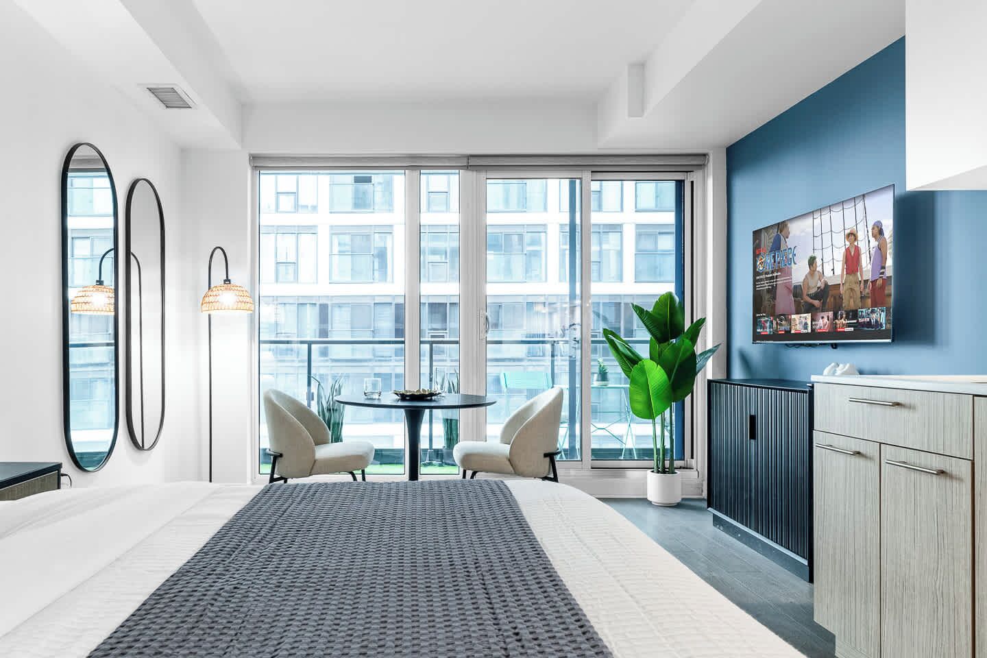A modern hotel room with a large window, a dining area with two chairs, a round table, a potted plant, and a TV mounted on the wall.