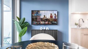 Una sala de estar moderna con televisor mostrando un programa, una mesa de comedor con una planta y una zona de cocina con un fregadero.