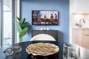 Ein modernes Wohnzimmer mit Fernseher, der eine Sendung zeigt, einem Esstisch mit Pflanze und einer Küchenzeile mit Spüle.
