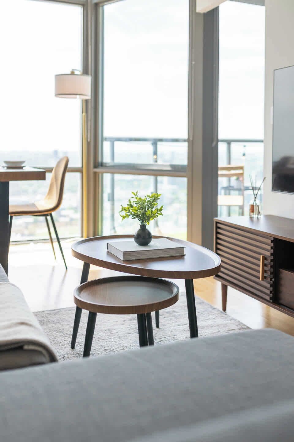 Een moderne woonkamer met een bank, een salontafel, een bijzettafeltje en een boekenkast. Een groot raam biedt uitzicht op de stadsgezicht.