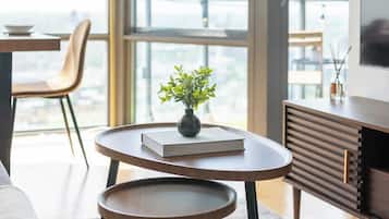 Un salon moderne comprenant un canapé, une table basse, une petite table d’appoint et une bibliothèque. Une grande fenêtre offre une vue sur le paysage urbain.